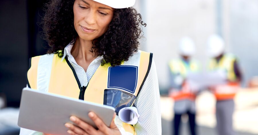 Female building engineer using a tablet on her work site Young female construction engineer wearing a safety vest standing outdoors on a worksite using a digital tablet with workers in the background